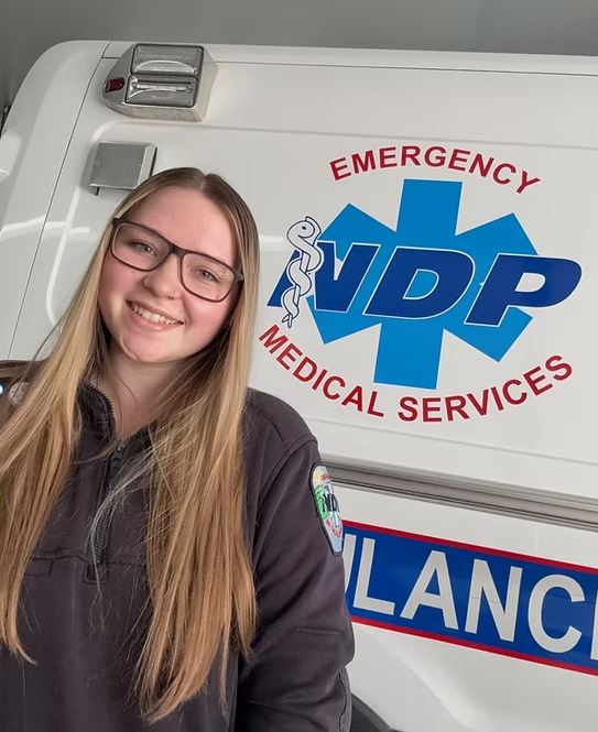 A young woman EMT stands in front of an ambulance.
